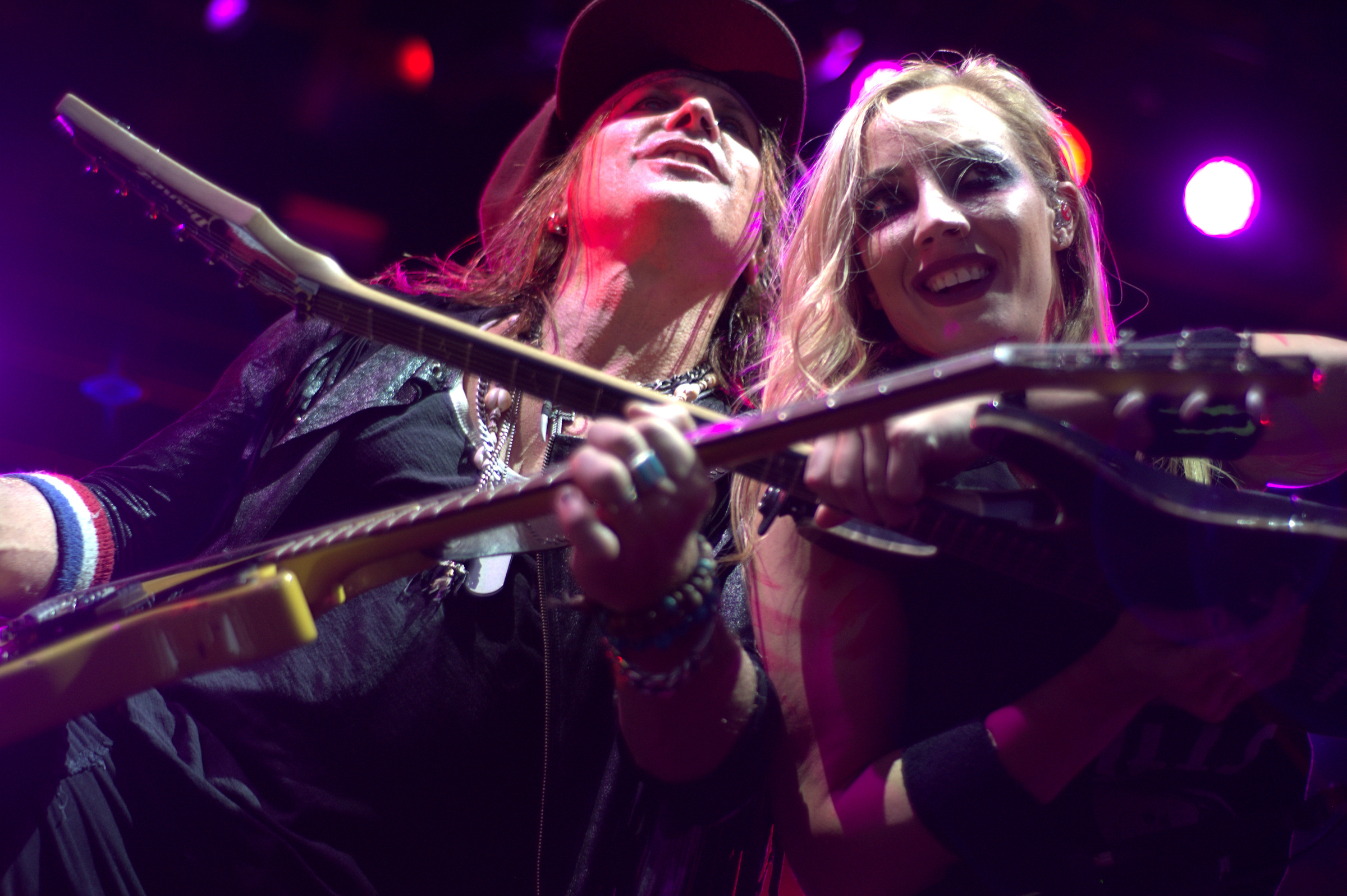 Two guitarists perform with their instruments crossed beneath purple stage lights.